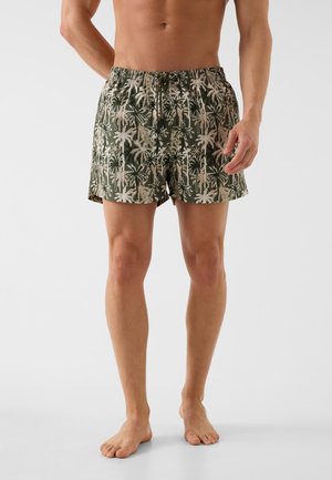 Męski tułów i nogi w zielonych kąpielówkach z beżowym nadrukiem palm, stojący boso na białym tle.