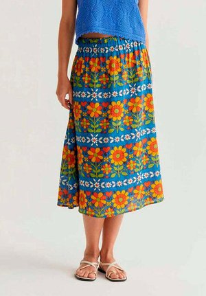 Mujer con falda midi azul con flores en colores naranja brillante, amarillo y blanco, combinada con sandalias beige de tiras.