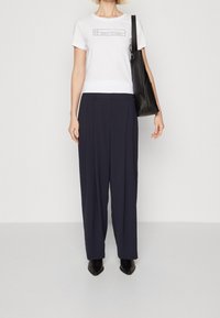 Valkoinen t-paita mustalla logolla; tummansiniset korkeavyötäröiset housut, joissa on etupuolelle taitoksia; musta olkalaukku; mustat nilkkurit. Yksinkertainen tausta.