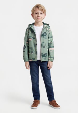 Gutt med blondt hår som har på seg en grønn jakke med byggeplasskjøretøytrykk, hvit t-skjorte, blå jeans og brune joggesko, stående mot hvit bakgrunn.