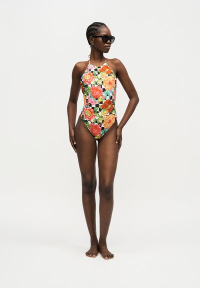 Kvinne iført en fargerik blomster mønstret halterneck badedrakt og svarte solbriller, stående barfot mot en ensfarget bakgrunn.