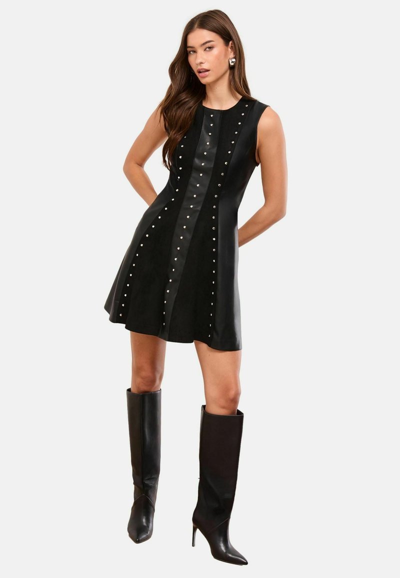 Černé šaty bez rukávů s rozšířenou sukní, s vertikálními koženými panely a stříbrnými nýty. Kombinované s černými vysokými lodičkami ke kolenům.