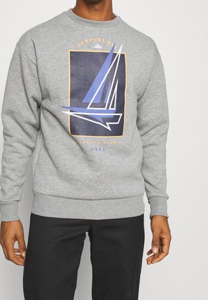 Mand iført grå sweatshirt med navy og hvid sejlbåds grafik og tekst, der lyder Newport Bay Sailing Club 1985, sammen med sorte bukser.