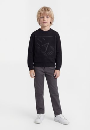 Blond deček stoji, oblečen v črno EA7 majico z dolgimi rokavi, sive hlače in bele superge na belem ozadju.