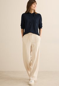 Mörkblå hoodie med dragsko och ribbade ärmslut, i kombination med krämfärgade, plisserade byxor och vita sneakers. Slät stickad textur genomgående.