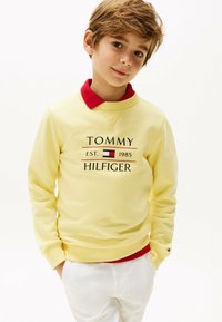 Sudadera amarilla con un cuello rojo, con un logo en texto negro: "TOMMY HILFIGER EST. 1985." Tejido suave y puños acanalados.