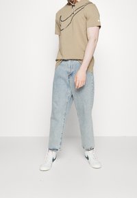 Béžové tričko s černým logem Nike, v kombinaci s světle modrými džíny rovného střihu a bílými teniskami s zeleným akcentem.