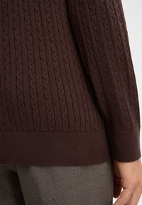 Brauner Strickpullover mit einem Zopfmuster, ribbeligem Saum und Bündchen, mit einer strukturierten Oberfläche. Betont durch enganliegende Ärmel.