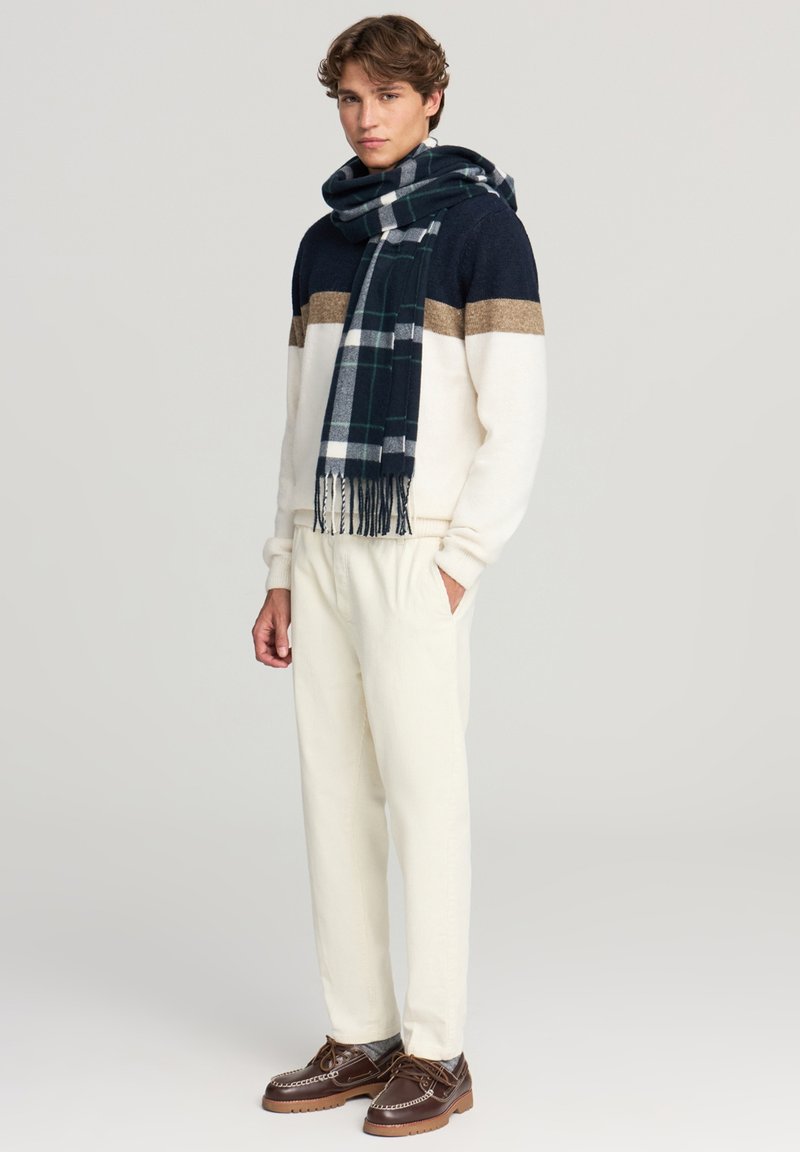 Mężczyzna ubrany w granatowy, brązowy i biały sweter, kremowe spodnie, brązowe skórzane buty i granatowy szalik w kratę, stoi z rękami w kieszeniach.