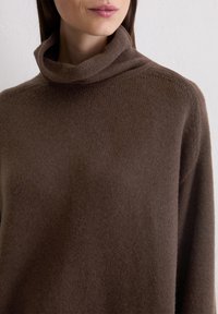 Brązowy sweter z wysokim dekoltem, wykonany z miękkiego, teksturowanego materiału; luźny krój z długimi rękawami. Brak widocznych wzorów i dodatków.