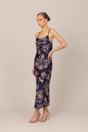 Mujer de pie con un vestido de tirantes finos floral en azul marino, pendientes dorados, pulsera y sandalias de tacón beige sobre un fondo beige.