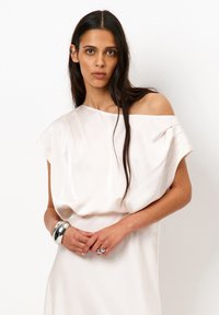 Lichtbeige satijnen jurk met een off-shoulder ontwerp, zachte draping en korte mouwen. Zilveren metalen armbanden voegen detail toe.