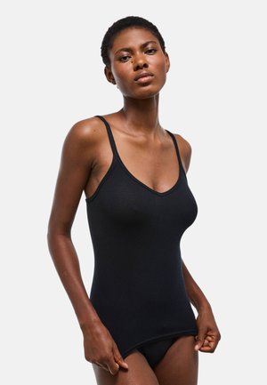 Femme portant un débardeur côtelé noir ajusté et des sous-vêtements noirs assortis sur un fond clair uni, posant avec une main tirant sur le haut.