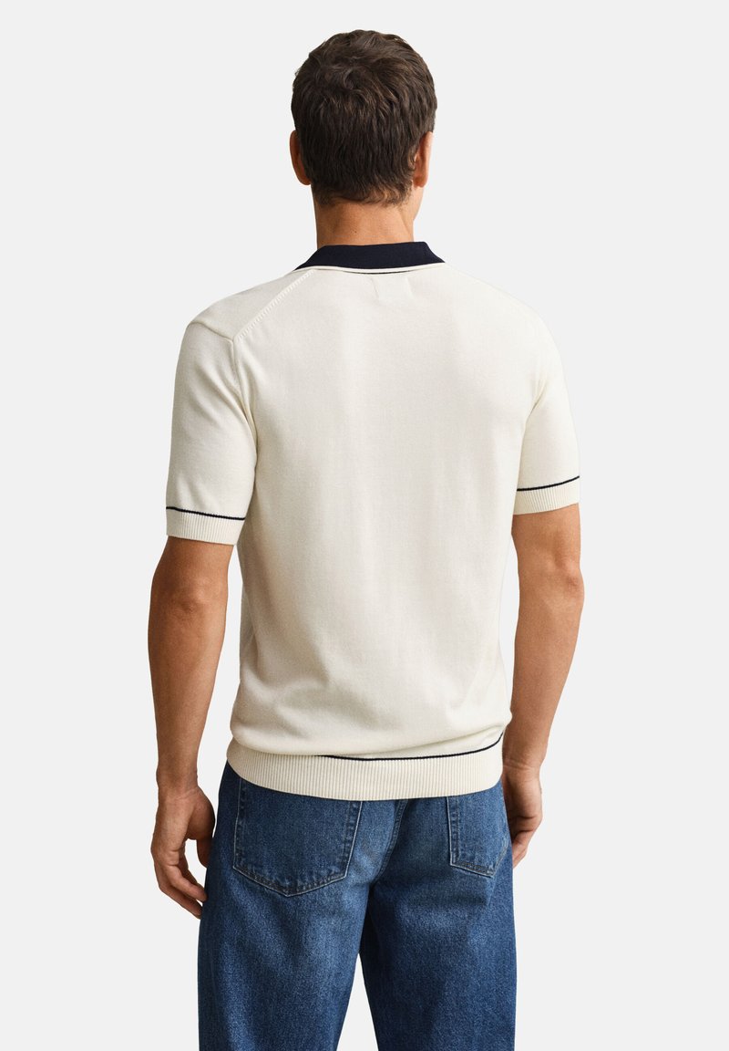 Uomo di spalle che indossa un maglione crema a maniche corte con colletto e bordi neri, abbinato a jeans di denim blu.