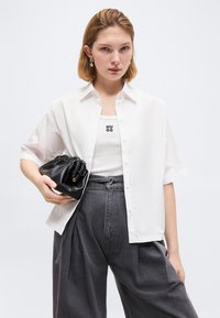 Νέα γυναίκα που φοράει άσπρο πουκάμισο με κοντά μανίκια πάνω από ένα λευκό τοπ Hugo, γκρι ψηλόμεσα παντελόνια με φαρδιά φάσα, κρατώντας μια μαύρη τσάντα clutch με πτυχώσεις.