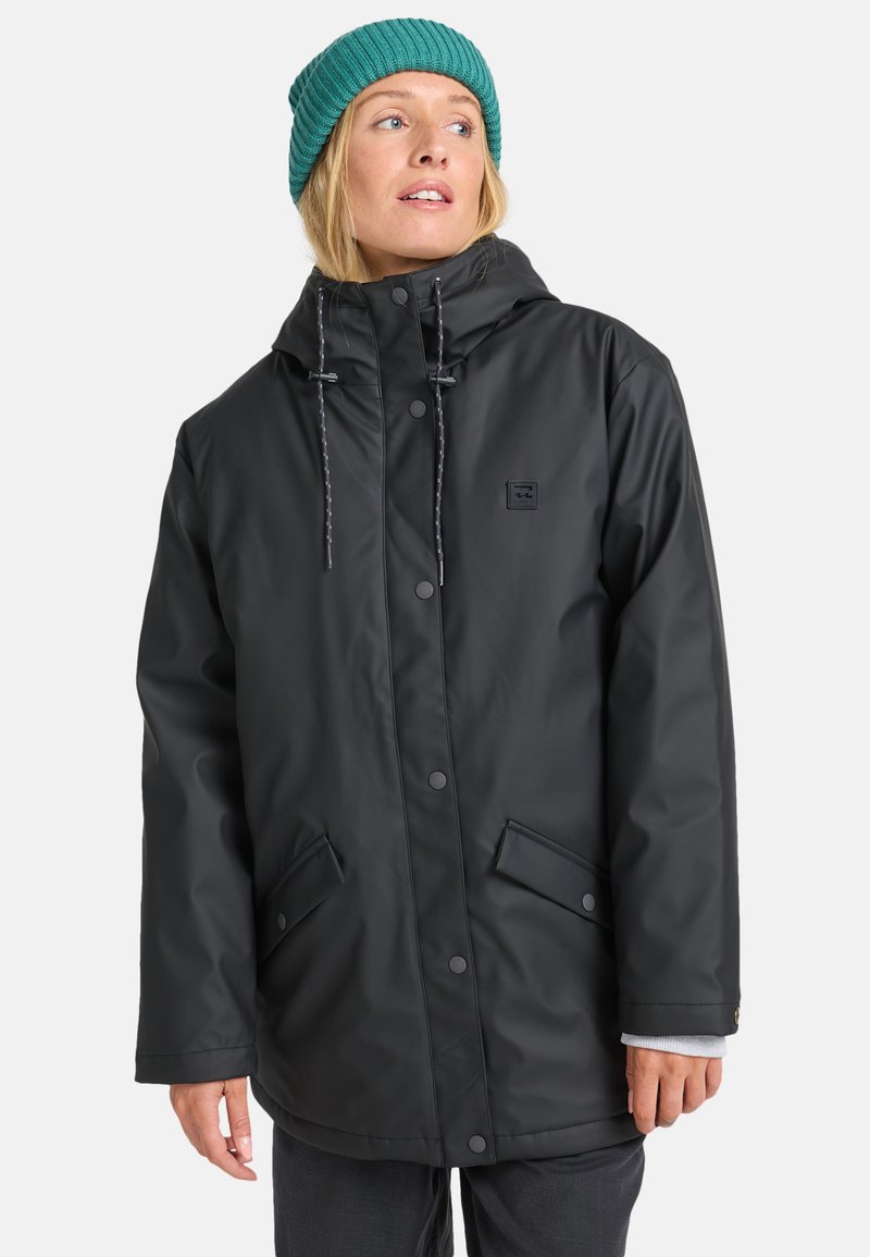 Chaqueta negra impermeable con capucha, que incluye botones a presión, cordones ajustables y dos bolsillos frontales. Se lleva con un gorro de punto color teal.