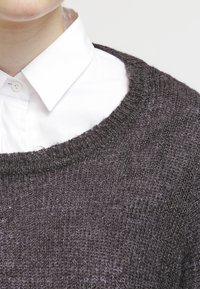 Tummanharmaa neulottu villapaita, leveällä pyöreällä kaula-aukolla, teksturoidulla pinnalla, valkoisen kauluspaitapuseron päällä, jossa näkyy nappidetalji.