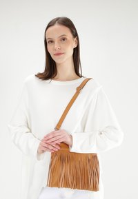 Torebka crossbody z brązowej skóry z frędzlami, z regulowanym paskiem i gładką fakturą. Noszona na białej bluzce na neutralnym tle.