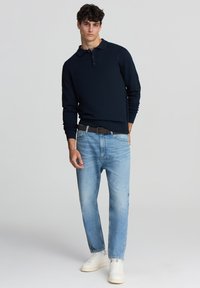 Polo a maniche lunghe blu navy con colletto e tre bottoni. Jeans dritti blu chiaro, indossati con sneakers bianche e una cintura marrone.