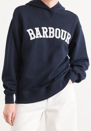 Personne portant un sweat à capuche Barbour bleu marine et un pantalon blanc, une main dans la poche et l'autre détendue sur le côté.