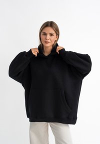 Felpa nera oversized realizzata in tessuto morbido, con tasca frontale, maniche ampie e cappuccio regolabile. Texture liscia, design minimale.