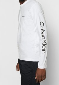 Person in einem weißen Langarmshirt mit "Calvin Klein" auf der Brust und dem Ärmel, kombiniert mit schwarzen Hosen, steht vor einem weißen Hintergrund.