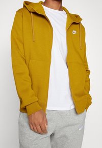 Hombre vestido con una sudadera con cremallera color mostaza, una camiseta blanca debajo y pantalones de chándal gris claro con logotipos de Nike en la sudadera y los pantalones.