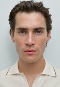 Ritratto di una persona con capelli corti e mossi di colore marrone e occhi blu. Indossano una maglietta polo beige chiaro con colletto a punta e bordature scure.