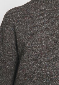 Grauer Strickpullover mit strukturiertem Stoff und farbigen Sprenkeln, die auf dunklem Hintergrund einen mehrfarbigen Effekt erzeugen.