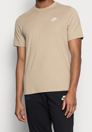 Beiges kurzärmeliges Nike T-Shirt, getragen von einer Person, kombiniert mit schwarzen Nike Jogginghosen mit Logo und Schriftzug am linken Bein.