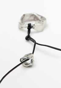 Anneau en métal argenté de forme irrégulière, relié à un cordon en cuir noir. Les détails notables comprennent une surface lisse et réfléchissante.