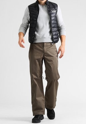 Homme portant un gilet matelassé noir, une chemise grise à manches longues, un pantalon marron et des chaussures noires marchant sur un fond blanc uni.