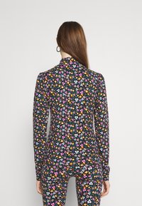 Frau mit langen braunen Haaren, die ein schwarzes, floral gemustertes Langarmoberteil und Hose trägt, steht mit dem Rücken zu einer schlichten Hintergrund.