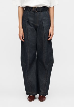 Persoon die hoge taille, wijde donkerdenim broek draagt met een zwarte riem en bruine schoenen, staand tegen een effen achtergrond.