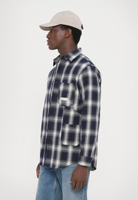 Marineblå og hvit rutete flanellskjorte med lange ermer, lomme detalj, og kragehals, kombinert med lyseblå denim jeans og en beige caps.