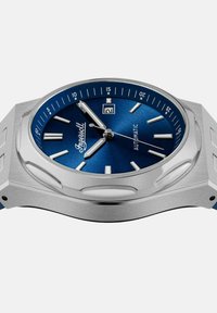 Zegarek ze stali nierdzewnej z niebieską tarczą, srebrnymi znacznikami godzin i wyświetlaczem daty i dnia miesiąca. Charakteryzuje się kanciastą formą koperty i polerowanymi akcentami.