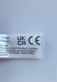 White fabric tag with black text. Displays UK and CE certification marks, brand name, address, and eco-friendly production note.