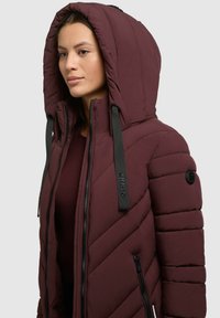 Burgundijas pūkainais mētelis ar lielu kapuci, izteiktu quilted tekstūru, rāvējslēdzējiem un kontrastējošām melnām siksnām ar zīmola akcentiem.