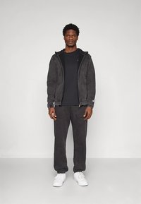 Čierna textúrovaná mikina na zips, v kombinácii s čiernym tričkom a zodpovedajúcimi teplákmi. Oblečenie dopĺňajú biele tenisky.