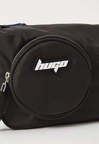 Borsa in tessuto nero con una tasca rotonda con zip, caratterizzata da un logo "hugo" bianco. Texture liscia e dettagli cuciti lungo i bordi.