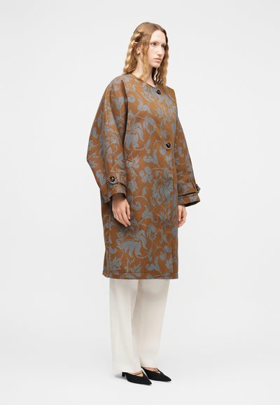 Γυναίκα με κυματιστά ξανθά μαλλιά, ντυμένη με καφέ παλτό με λουλουδάτο μοτίβο, λευκό παντελόνι φαρδιάς γραμμής και μαύρα μυτερά παπούτσια, στέκεται μπροστά σε λευκό φόντο.