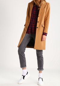 Manteau long en camel avec un design ajusté, devant à une seule boutonnière, associé à une chemise à carreaux, un jean gris et des chaussures blanches à enfiler.