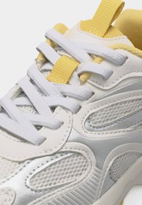 Λευκό και ασημί αθλητικό παπούτσι με κίτρινες λεπτομέρειες, λευκά κορδόνια, διχτυωτά πάνελ και κίτρινη θηλιά έλξης στη γλώσσα.