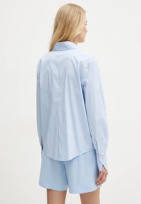 Camicia azzurra chiara con colletto e maniche lunghe, realizzata in tessuto testurizzato, con spacchi laterali e polsini abbottonati, indossata con pantaloni corti coordinati.