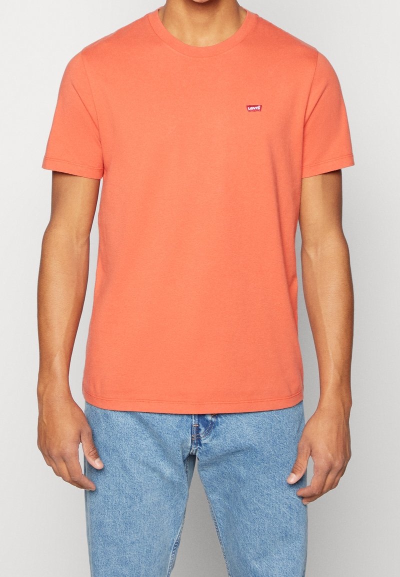Homme portant un t-shirt Levi's corail à manches courtes et un jean bleu clair, debout devant un fond uni clair.