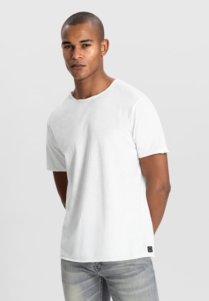 Weißes T-Shirt mit Rundhalsausschnitt und kurzen Ärmeln, aus leichtem Stoff gefertigt. Besonders ist ein kleines Logoschildchen in der linken unteren Ecke.