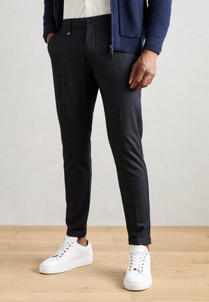 Calças acinzentadas escuras, ajustadas, com um padrão discreto, apresentando um fecho frontal com botão e bolsos laterais, combinadas com ténis brancos.