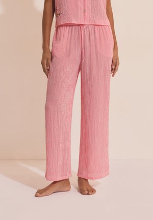 Femme portant un pantalon ample à rayures verticales roses et blanches avec une taille à cordon, debout pieds nus sur un fond beige.