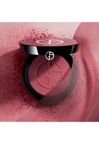 Rund blush kompakt med et glanset svart lokk som har en logo. Inneholder et mykt, matt rosa pudder, med løse pigmenter spredt rundt.