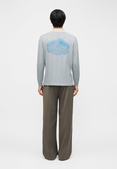 Άτομο με κοντά μαύρα μαλλιά φοράει ανοιχτό γκρι πουλόβερ με μπλε μοτίβο σύννεφου σε σχέδιο δίχτυ στο πίσω μέρος και χαλαρά καφέ παντελόνια, στέκεται με την πλάτη.